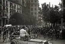 Photographie en noir et blanc d'un peloton de coureurs roulant en ville, acclamés par la foule.