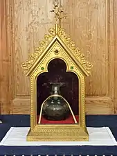 Photographie en couleurs d'une fiole en verre décorée d'un ruban dans un reliquaire en cuivre vitré.