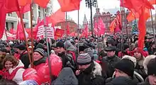 Photo couleur de manifestants défilant dans une rue et portant des drapeaux rouges.