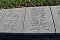 Une rangée de pierres rectangulaires posées à plat sur le sol. Y est gravée une liste de noms de soldats allemands, leur grade ou poste et leur date de mort.