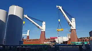 À l'arrière plan, la grue du navire de transport vient de déposer la turbine sur son ber. La grue est encore équipée du palonnier. Au premier plan de gauche à droite on voit des sections des piles des futurs éoliennes, et des bers vide pour en accueillir de nouveaux. Au premier plan au milieu à gauche on distingue une plateforme automotrice (verte) qui sert à déplacer les éléments, et à droite à l'arrière plan une des grues mobiles (verte) qui permet de manipuler les éléments... Cette grue mobile n'est pas suffisante pour déplacer les piles ou les turbines, une grande structure porteuse (non photographiée ici) sert à redresser les piles, qui arrivent horizontales par bateau, et sont redressées avant d'être stockées verticales.