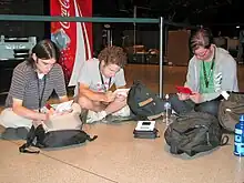 Trois jeunes joueurs de jeux vidéo sont assis sur le sol, absorbés par le jeu sur leur console portable.