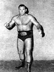 Photo en noir et blanc d'un catcheur brun torse nu. Il porte un slip noir et une fine ceinture.