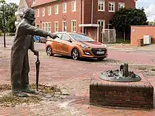 Photographie en couleur montrant une statue d'un vieil homme appuyé sur un parapluie et pointant le doigt vers une ville miniature en ruine.