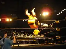 Un catcheur effectue une prise du haut de la troisième corde en portant son adversaire sur ses épaules. Ils sont en l'air et s'apprêtent à passer à travers un panneau de bois recouvert de fils barbelés.