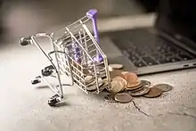 Près d’un ordinateur portable, un panier d’achats sur roues miniature est renversé et déverse des sous.