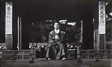 Photographie en noir et blanc montrant un homme assis sous un auvent en bois et vêtu d’une robe richement brodée de dragons sur la poitrine et les épaules.