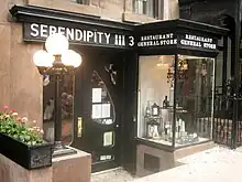 Photographie d'une devanture vitrée avec enseigne brun foncé, bow-window sur la droite de l'image, lampadaire et jardinière de fleurs sur la gauche de l'image.