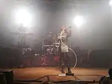 Trois membres d'un groupe sur une scène. La chanteuse tient un trépied en forme de S