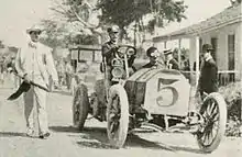 Photo d'une voiture à l'arrêt, avec son pilote et son mécanicien embarqué, ainsi qu'un officiel de course, à côté.