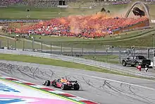A matte navy single-seater open-wheel racing drives around a corner on a tarmac course while a grandstand full of people dressed in orange set off orange fireworks in the background.