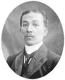 Photo en noir et blanc. Buste d'un homme dans la trentaine, fine moustache, costume et cravate, cheveux courts peignées en arrière