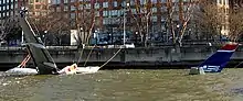 Photo montrant l'avion au premier plan amarré à une jetée, partiellement submergé dans le fleuve Hudson. Il est relié par des câbles à la berge et seule sa queue et son aile gauche sont clairement visibles, le reste de son corps se trouvant sous l'eau.