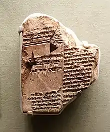 Photographie d'une tablette  fragmentée en argile beige couverte d'écritures cunéiformes anciennes. La tablette est inégale et incomplète, avec des bords cassés. Elle montre des signes d'érosion qui témoignent de son âge et de son origine historique. Les lignes de texte cunéiforme sont incomplètes.