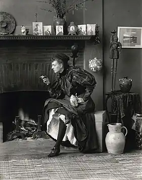 Autoportrait d'une femme assise devant une cheminée, tournée vers la gauche, tenant une cigarette dans une main et une bière dans l'autre.