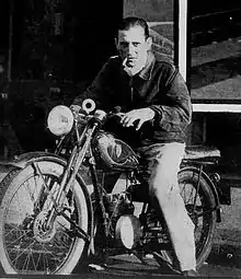 Photographie en noir et blanc. Un homme est assis sur une moto. Il porte un pantalon clair et une veste foncée. Son visage et vu de face et il a une cigarette à la bouche. La moto est orientée vers la gauche de l'image.