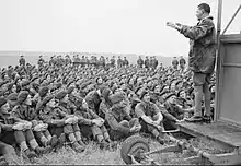 Photo en noir et blanc d'un homme en uniforme s'adressant depuis une estrade à une foule d'autres hommes assis.