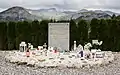 Photo d'une plaque rectangulaire grise, mesurant environ un mètre de haut et comportant des inscriptions dorées. Divers objets religieux et photos se trouvent autour. À l'arrière-plan se trouvent de petits arbustes de même hauteur que la stèle et, plus haut, une chaîne de montagnes.