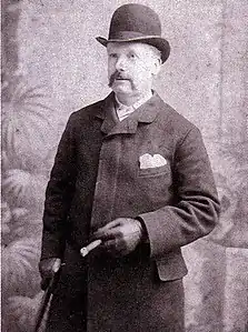 Photographie en noir et blanc. Homme au chapeau melon, avec manteau et gants, une canne sous le bras droit.