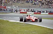 Photographie d'une monoplace F1 rouge, portant le numéro 28, vue de trois-quarts, proche de l'herbe.