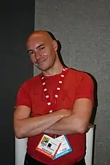 Photo d'un homme chauve souriant portant un haut à manches courtes rouge et des badges d'accès autour du cou.