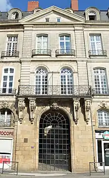 Façade ensoleillée de pierre grise, surmontée du fronton. Le toit en ardoise noire et les cheminées en briques rouges se détachent sur fond de ciel bleu.