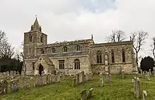 Photo d'un long bâtiment en pierres ocres flanqué d'une haute tour du côté gauche, au milieu d'un pré vert jonché de pierres tombales
