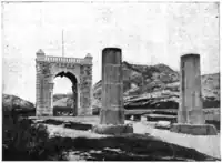 Photographie en noir et blanc montrant à l’avant-plan deux piliers en bois et un arc de triomphe en pierre à l’arrière-plan.