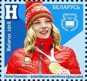 Timbre avec un fond bleu représentant une femme blonde vêtue d'un bonnet et d'un manteau principalement rouge et tenant une médaille d'or accroché autour de son cou.