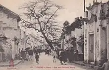 Photographie ancienne montrant des passants dans une rue composée de bâtiments de style extrême-oriental, visiblement mal entretenus.