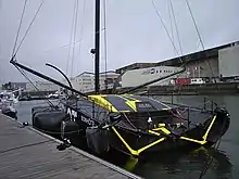 Vue de trois quarts arrière d'un voilier noir et jaune, qui a la particularité d'avoir un cockpit entièrement recouvert et fermé.