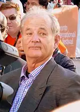 Photographie en buste d'un homme aux cheveux gris portant une chemise à carreaux et une veste grise.