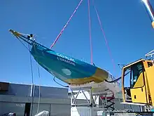 Vue en contre-plongée, depuis l'avant, du bateau entier posé sur son ber et suspendu à la grue.