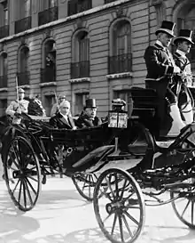 Photo en noir et blanc de deux individus moustachus et aux cheveux blancs, dans une calèche : l’homme situé à droite porte un haut-de-forme et regarde devant lui ; celui de gauche sourit au photographe ; à l’arrière-plan, deux gardes