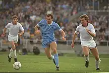 vue d'une rencontre de football, un joueur en bleu pousse le ballon poursuivi par un joueur en blanc