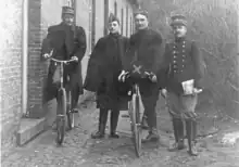 Photographie noir et blanc de quatre hommes en uniforme, deux sont sur des bicyclettes.