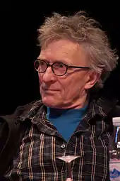 Homme de 59 ans, cheveux en bataille, lunettes rondes, col de chemise ouvert sur un tee-shirt bleu.