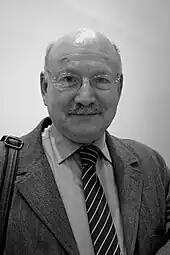 Un homme en costume, moustache, crâne dégarni et lunettes rondes, regarde devant lui.