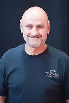 Photographie couleur d’un homme, de tronc, chauve, souriant, avec une barbe de 3 jours blanche et un t-shirt bleu marine