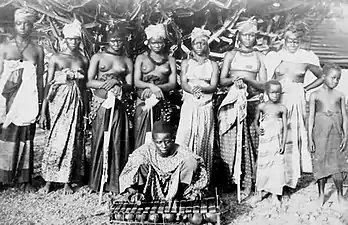 Photographie d'un groupe de femmes partiellement dénudées, fixant l'objectif. Un homme assis au premier plan joue d'un instrument de musique.