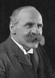 Portrait photographique en noir et blanc d'un homme portant une moustache grise très fournie.