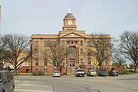 Vue de la façade d'un bâtiment de trois étages en bordure de route. Au premier plan des voitures sont visibles ainsi que des arbres sans feuillage.