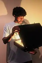 Un homme de race noire avec une coiffure afro vêtu comme le personnage de Jules Winnfield à la fin du film et ouvrant une mallette qui produit une forte lueur.