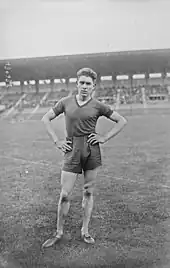 Photographie en noir et blanc d'un athlète, main sur les hanches, sur la pelouse au milieu d'un stade vide de spectateur.