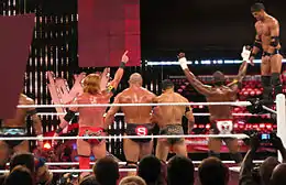 À droite, Justin Gabriel se tient debout sur la troisième corde du ring. Il s'apprête à effectuer sa prise de finition, le 450e splash, en se jetant sur Ricky Steamboat (non visible sur l'image). Les autres membres du Nexus, de dos, l'encouragent.