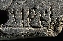 Photographie en couleurs d'une pierre rectangulaire gris foncé marquée de graphèmes, la partie gauche percée d'un trou, le coin en bas à droite présentant des stigmates de cassure.