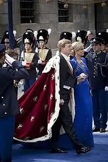 Un homme, portant un manteau d'hermine rouge, et une femme en robe bleue marche sur un tapis bleu pendant que deux hommes en uniforme leur rendent le salut militaire.