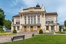Un bâtiment, grande façade au-dessus d'un escalier, deux statues sur son toit, autour un jardin avec une belle pelouse.