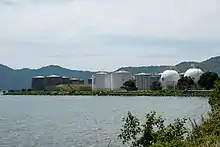 Photographie d'une usine pétrolière et de ses nombreuses citernes. L'ensemble est entouré par la mer au premier plan et par les montagnes à l'arrière-plan.