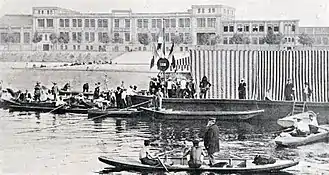 Photographie noir et blanc : des barques sur l'eau autour d'un ponton encombré de nombreuses personnes.